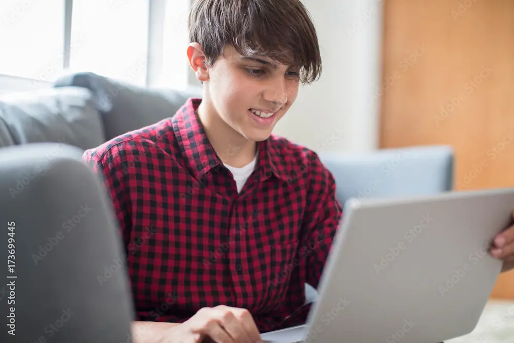 Teenager in a red plaid shirt using a laptop for telehealth sessions while sitting on a couch in a bright room.