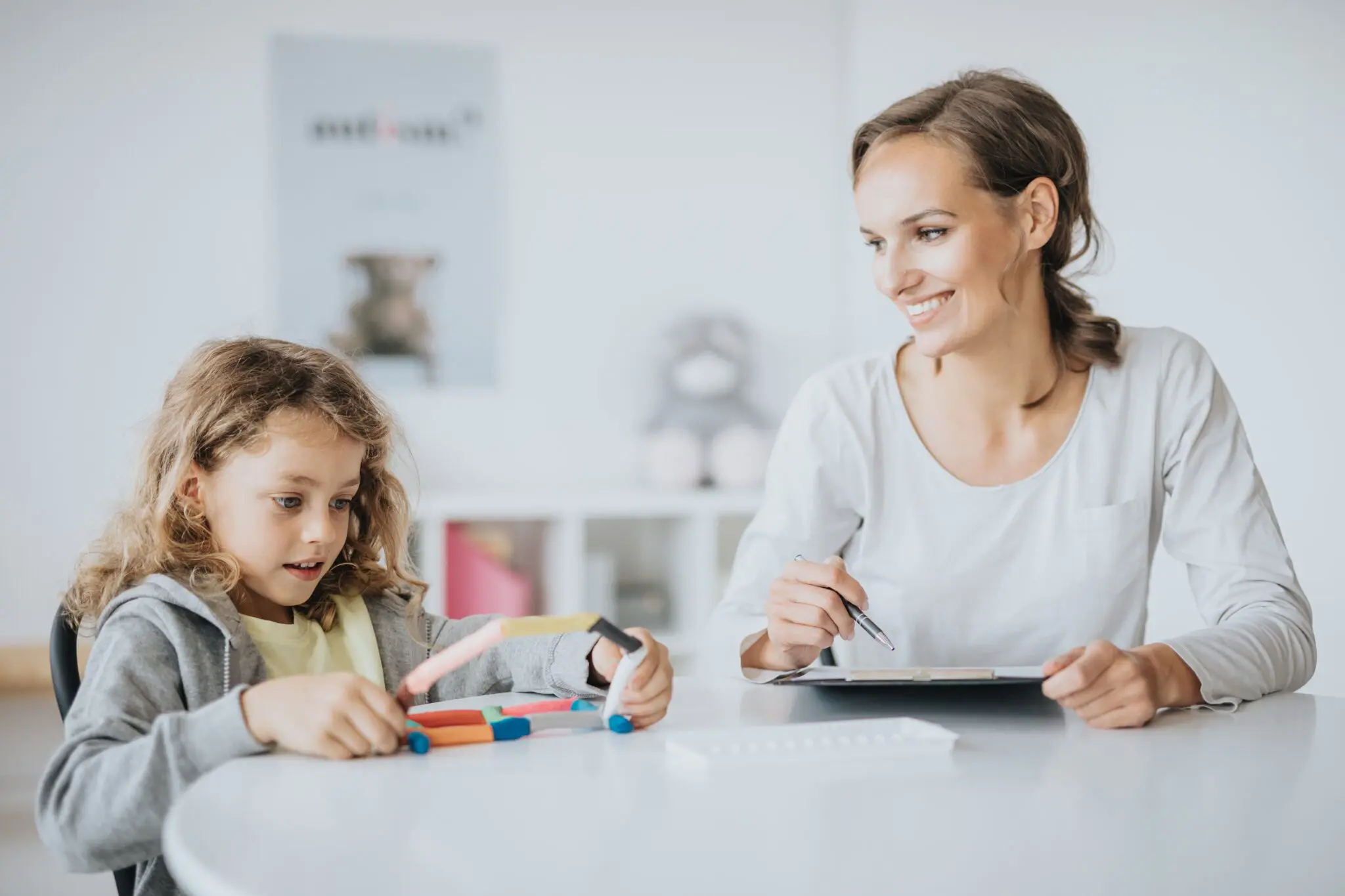 A woman and child sit at a table, the child playing with colored sticks while the woman, engaged in their telehealth autism therapy session, holds a pen and notepad, smiling warmly.