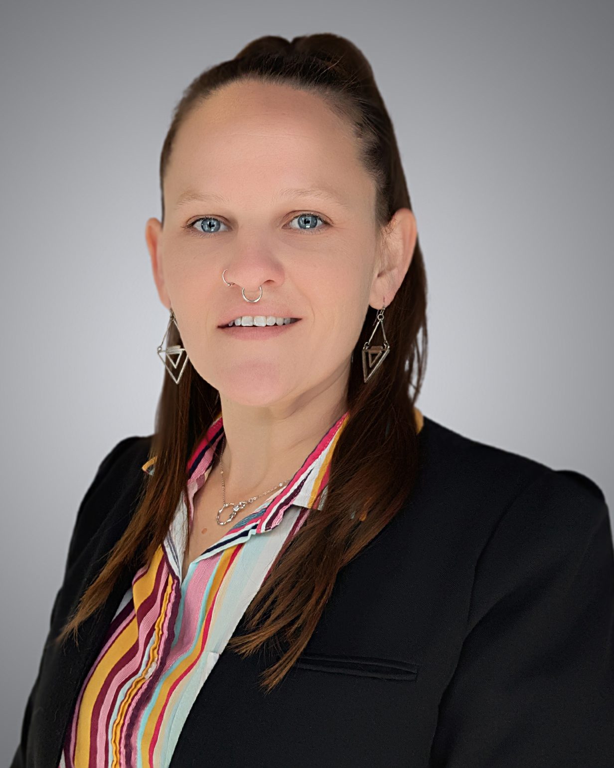 A woman with long brown hair in a half-up style, wearing a black blazer, striped shirt, geometric earrings, and a nose ring, poses against a plain gray background.