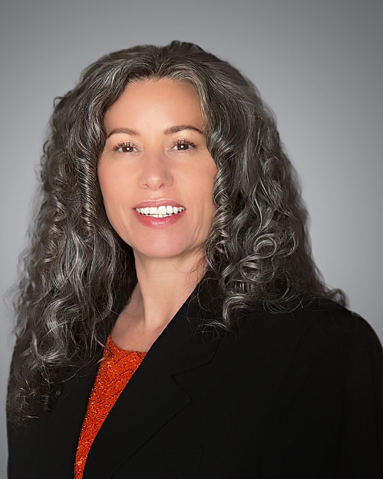 A woman with long, curly gray hair wearing a black blazer and an orange top, posed against a plain gray background.